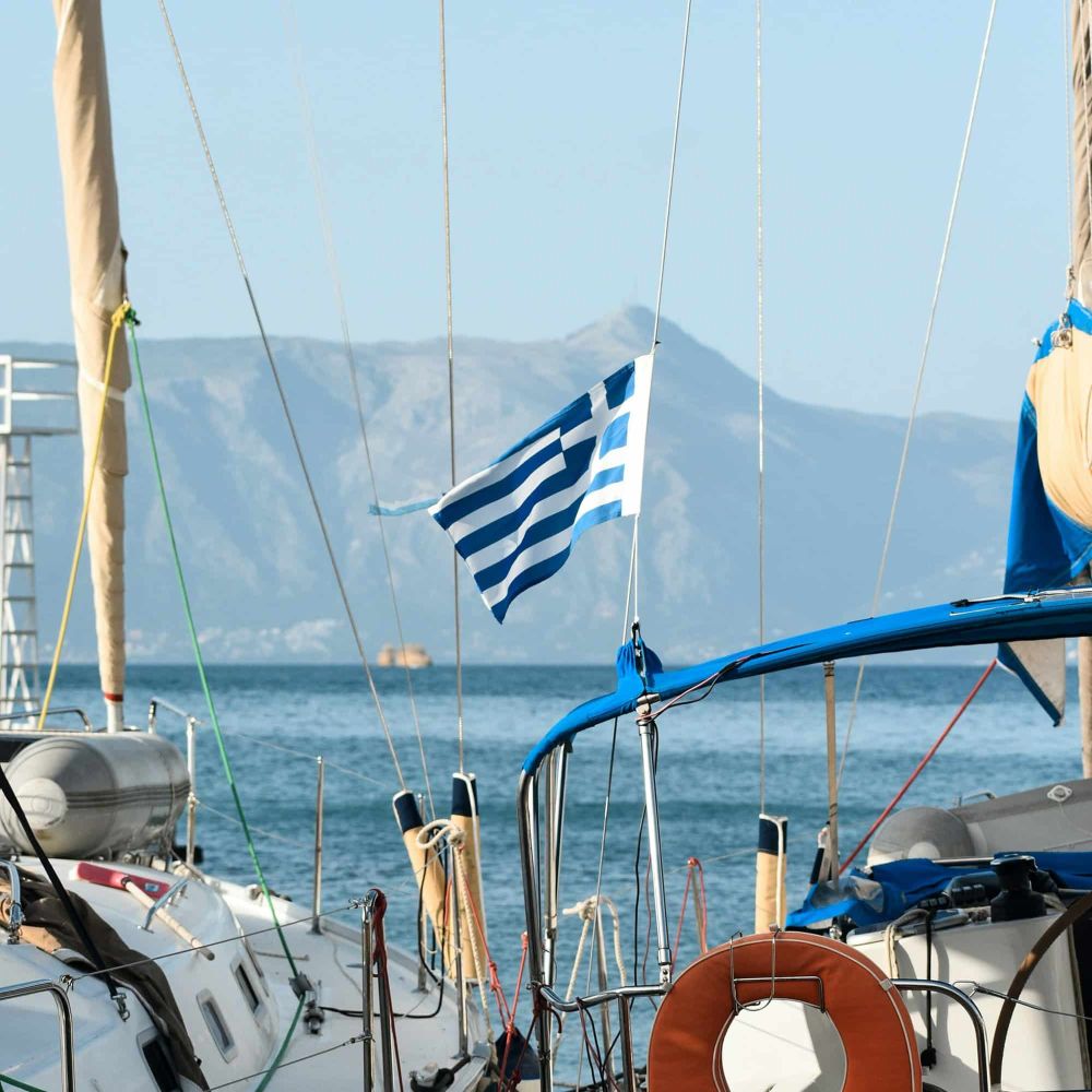 Zwei Segelboot mit einer Griechischen Flagge. Im Hintergrund der Ozean und ein Berg.